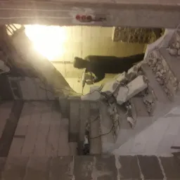 Pracownik w ciemnej bluzie wierci w betonowej konstrukcji schodów za pomocą wiertarki udarowej, widoczne gruz i fragmenty zniszczonych elementów, oświetlenie robocze w tle.