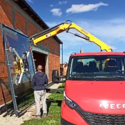 Montaż dużej szyby okiennej za pomocą przyssawkowej windy na samochodzie, widoczny budynek w trakcie budowy z czerwonej cegły i pole kukurydzy w tle.