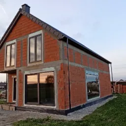 Surowy budynek z czerwonej cegły z zamontowanymi oknami z ciemnymi ramami, widoczne odbicie krajobrazu w szybach, teren wokół budynku w trakcie prac wykończeniowych.