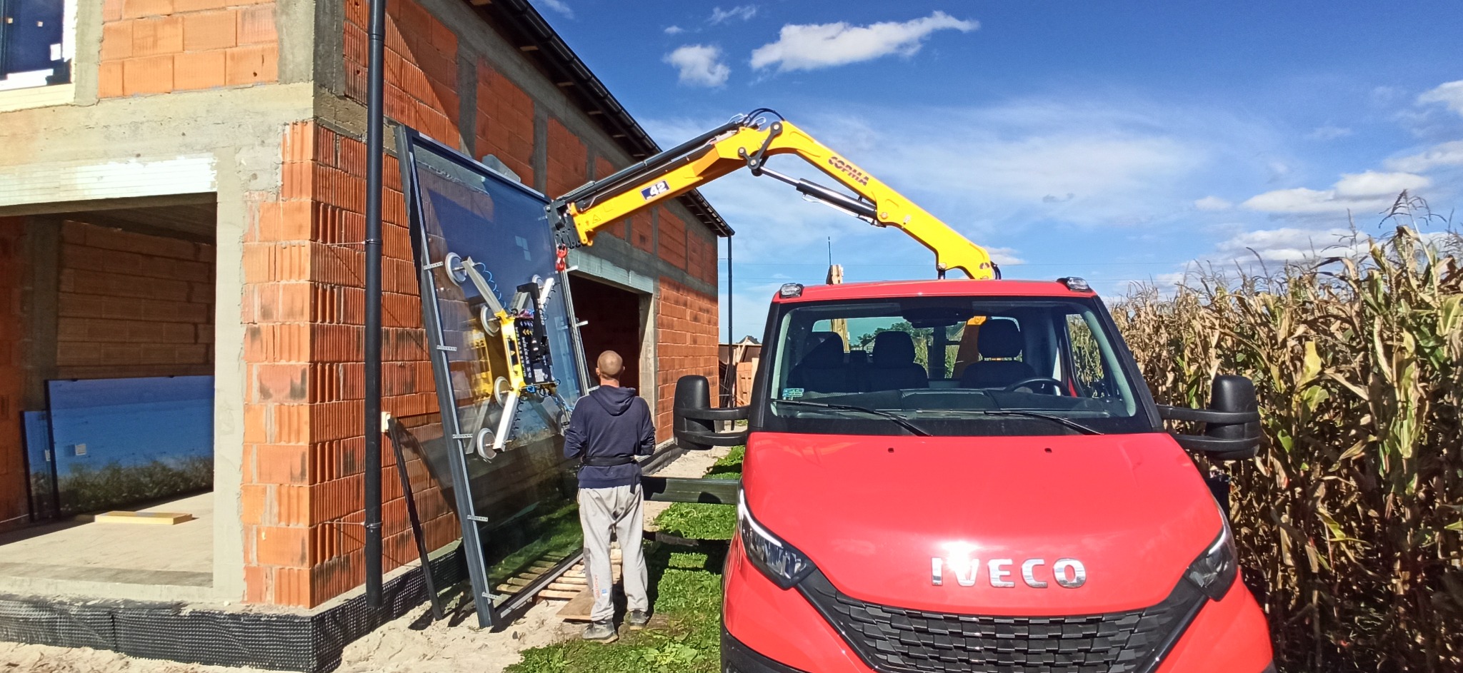 Montaż dużej szyby okiennej za pomocą przyssawkowej windy na samochodzie, widoczny budynek w trakcie budowy z czerwonej cegły i pole kukurydzy w tle.