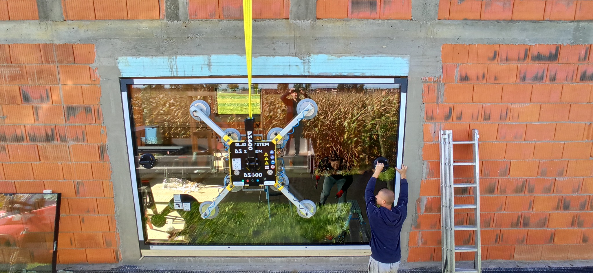 Montaż okna aluminiowego z użyciem przyssawki próżniowej, widoczny mur z cegły i drabina przystawna.