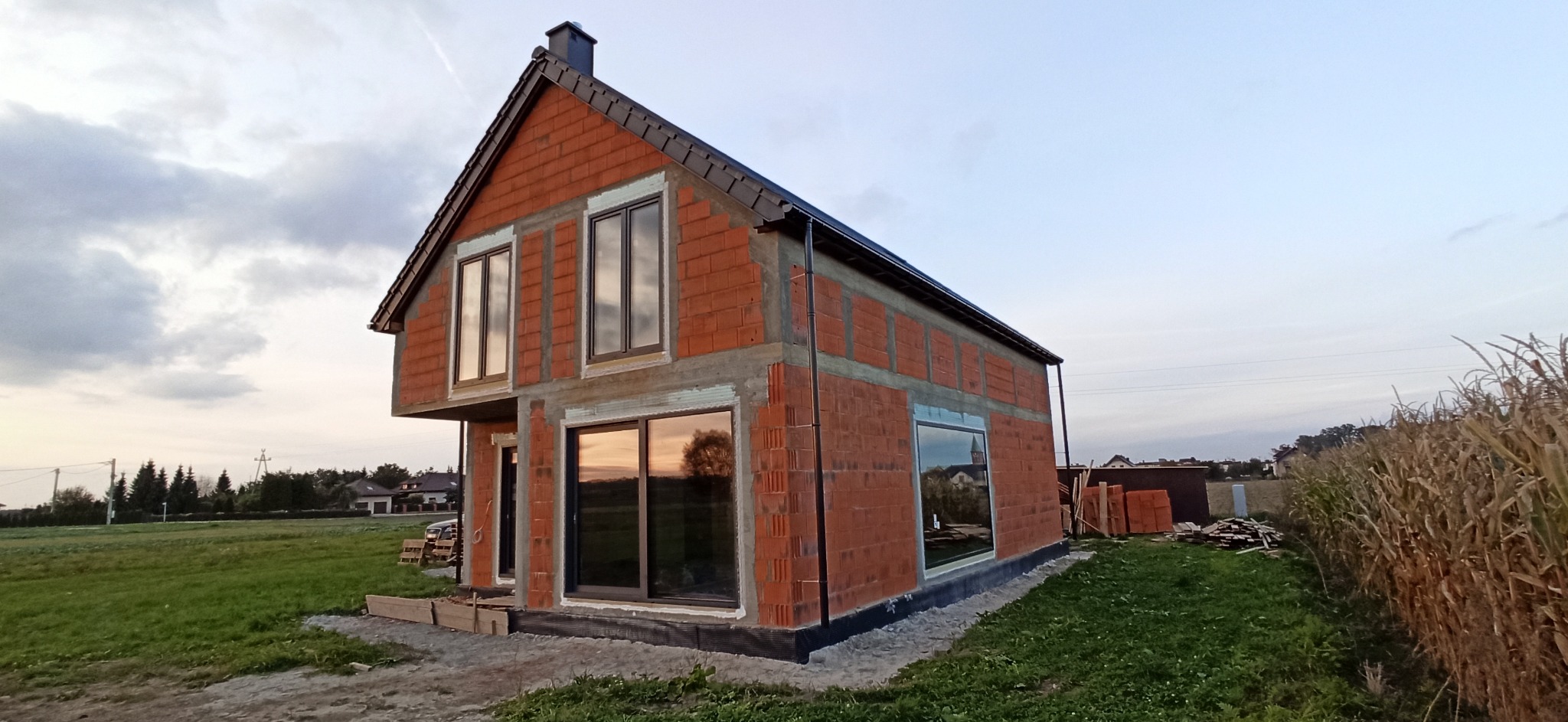 Surowy budynek z czerwonej cegły z zamontowanymi oknami z ciemnymi ramami, widoczne odbicie krajobrazu w szybach, teren wokół budynku w trakcie prac wykończeniowych.