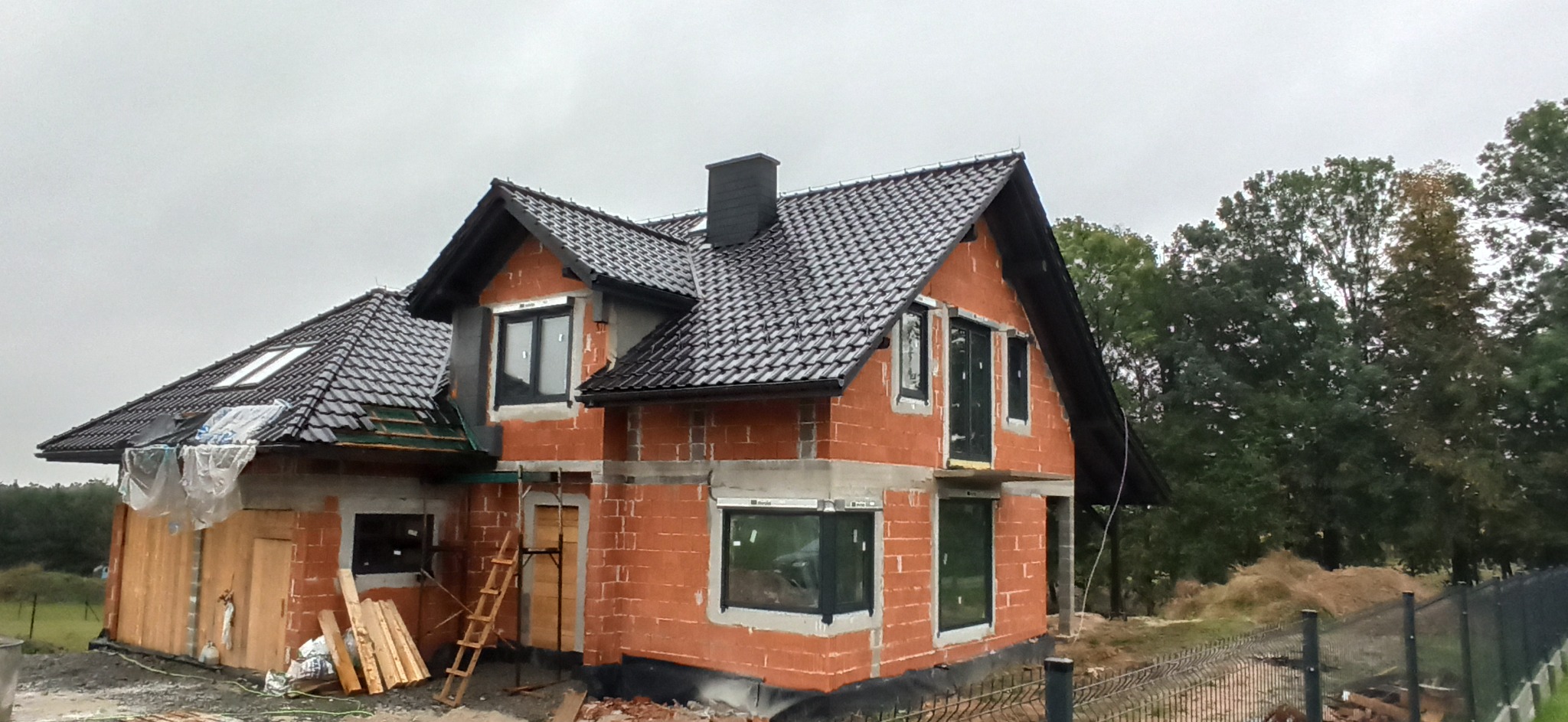 Budowa domu jednorodzinnego z czerwonej cegły, z zamontowanymi oknami aluminiowymi i czarną dachówką, widok z zewnątrz na plac budowy w pochmurny dzień.