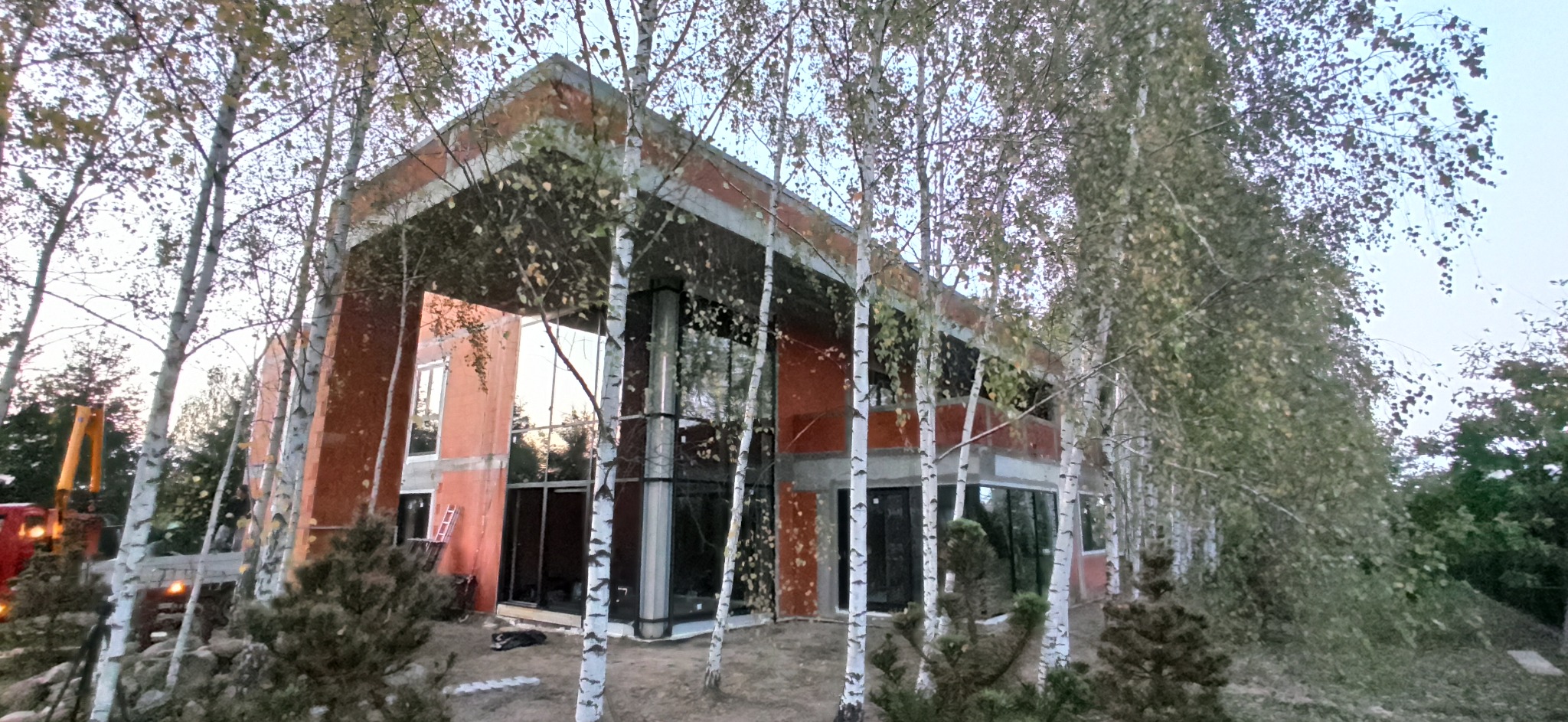 Nowoczesny dom w stanie surowym otwartym z dużymi przeszkleniami, widoczny zza brzozowego zagajnika, z dźwigiem budowlanym w tle.