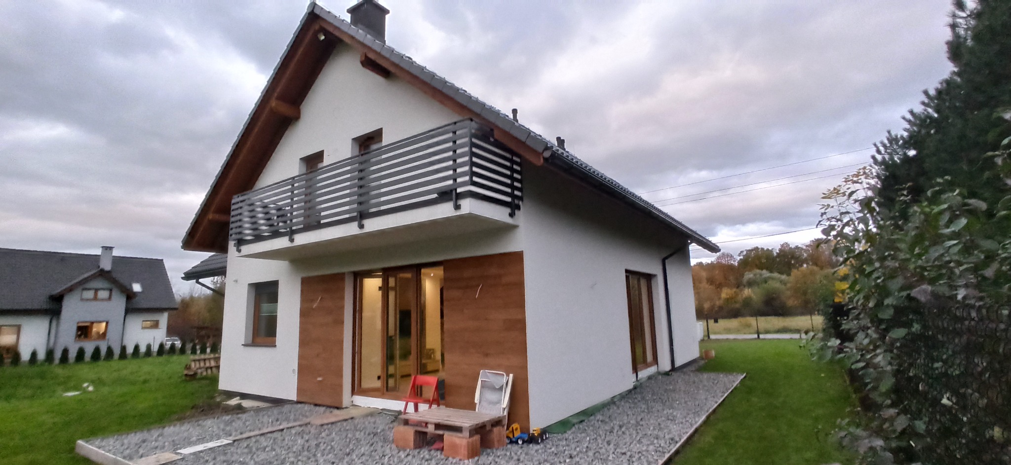 Nowoczesny dom z białymi ścianami, drewnianymi drzwiami przesuwnymi i balkonem z czarną balustradą, widok z zewnątrz na tle pochmurnego nieba.