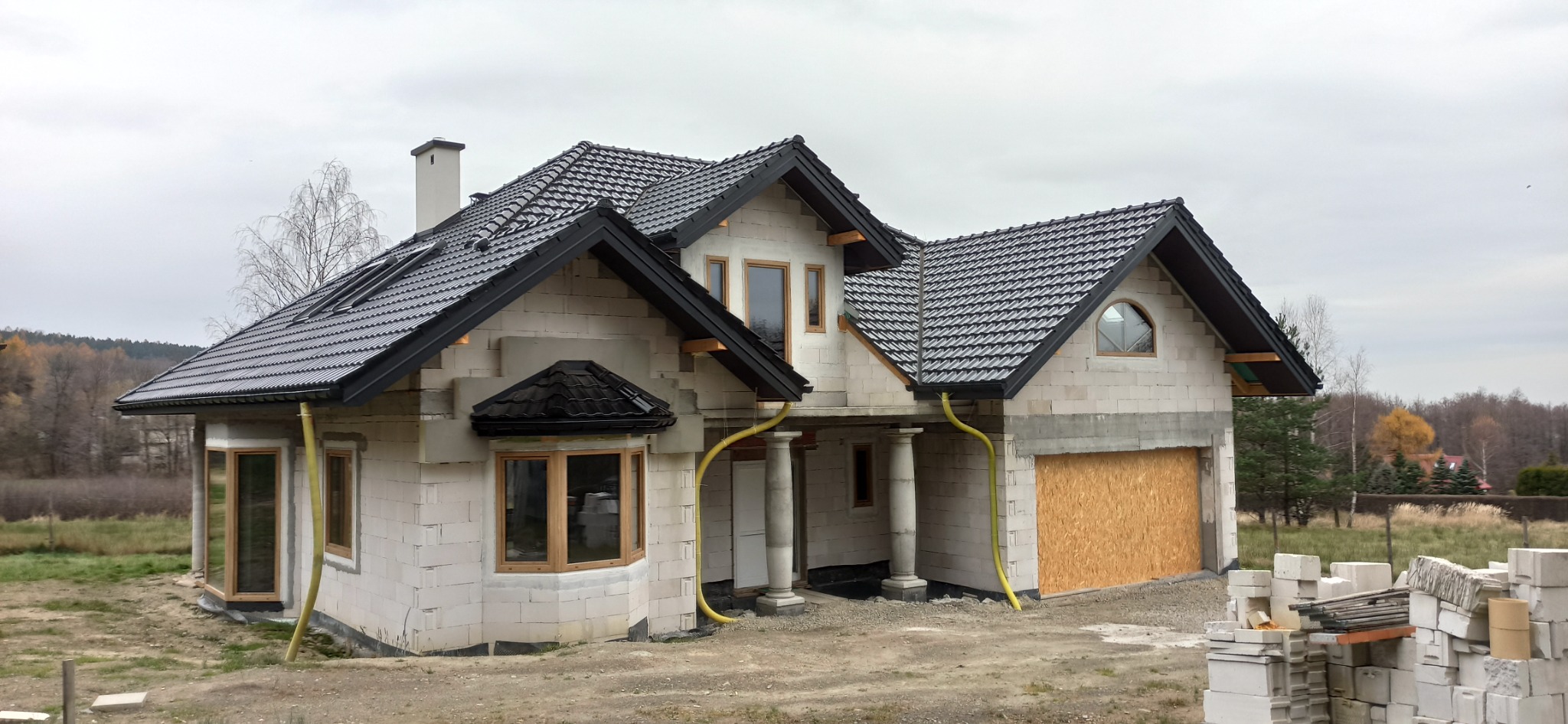 Surowy stan domu jednorodzinnego w trakcie budowy, widoczne czarne dachówki, jasne ściany z betonu komórkowego, drewniane ramy okienne i rury spustowe