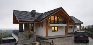 Nowoczesny dom jednorodzinny z drewnianą konstrukcją dachu i dużymi oknami, widoczny samochód na podjeździe z kostki brukowej.