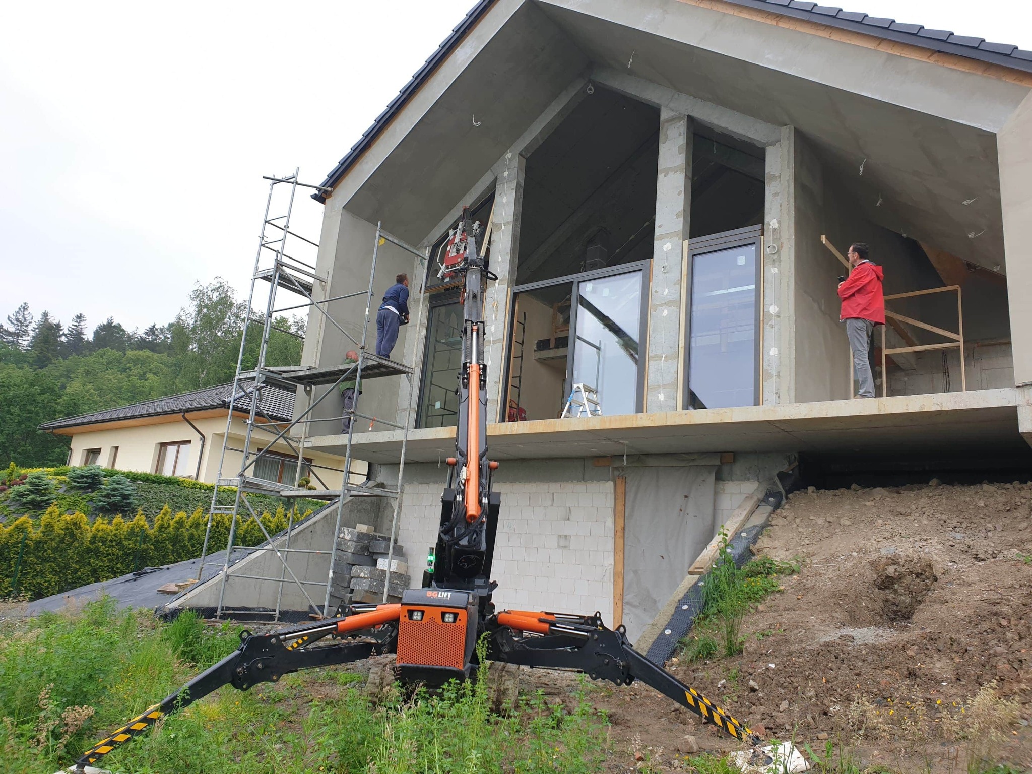 Montaż okien w surowym stanie budynku jednorodzinnego przy użyciu podnośnika teleskopowego; pracownicy na rusztowaniu i na podeście, widoczne okna z czarnymi ramami.