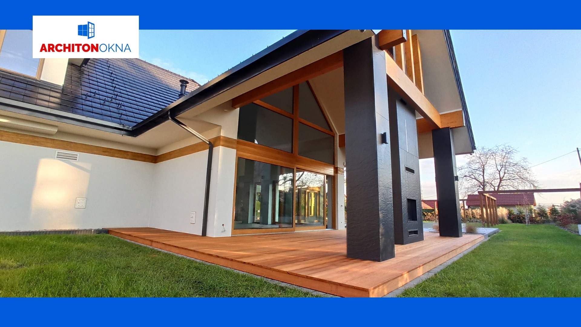 Nowoczesny dom z dużymi przesuwnymi drzwiami tarasowymi z drewnianą ramą, widok z zewnątrz na taras z desek, czarne kolumny podtrzymujące zadaszenie, trawnik i fragment ogrodu.