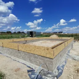 Fundamenty domu z bloczków betonowych, szalunek z płyty OSB, widoczna izolacja przeciwwilgociowa, w tle blaszany garaż i błękitne niebo z chmurami.