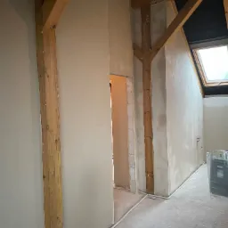 Ściana po tynkowaniu maszynowym w domu z drewnianą konstrukcją stropu i oknem dachowym. Widoczne świeże tynki, drewniane belki i fragment podłogi.