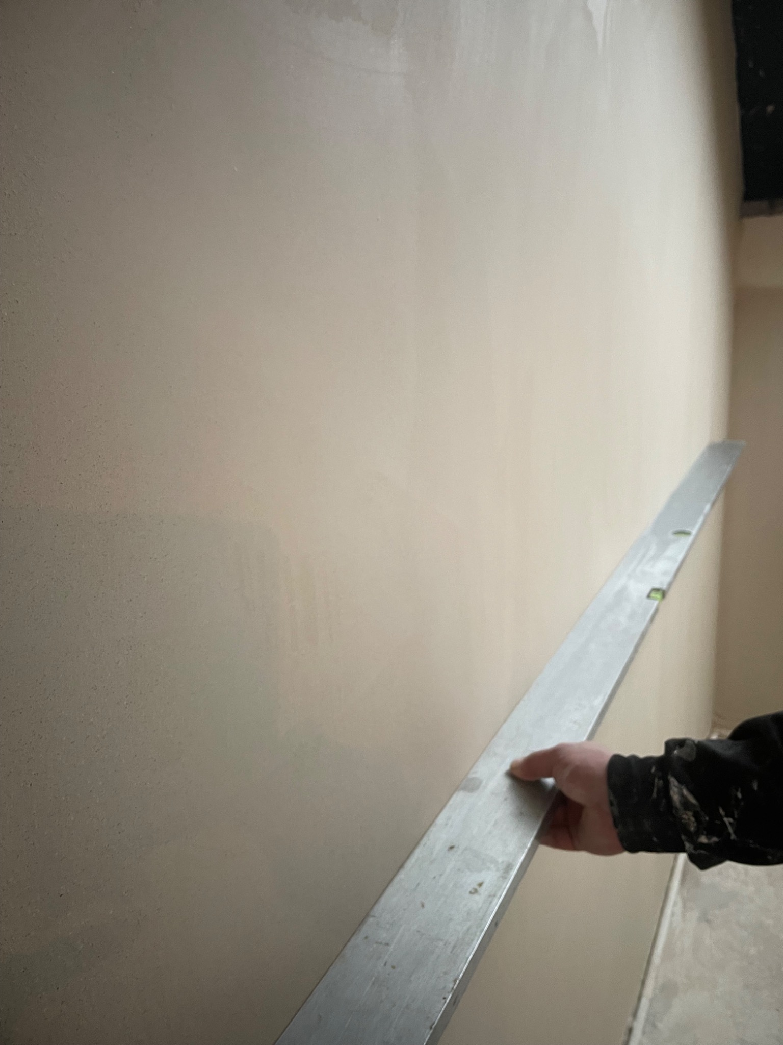 Świeżo nałożony tynk na ścianie wyrównywany długą łatą, w tle fragment sufitu. Detal z prac wykończeniowych.