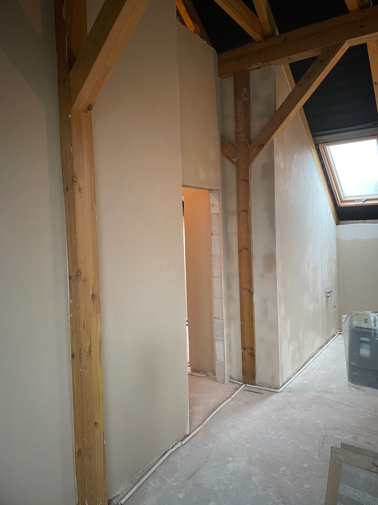 Ściana po tynkowaniu maszynowym w domu z drewnianą konstrukcją stropu i oknem dachowym. Widoczne świeże tynki, drewniane belki i fragment podłogi.
