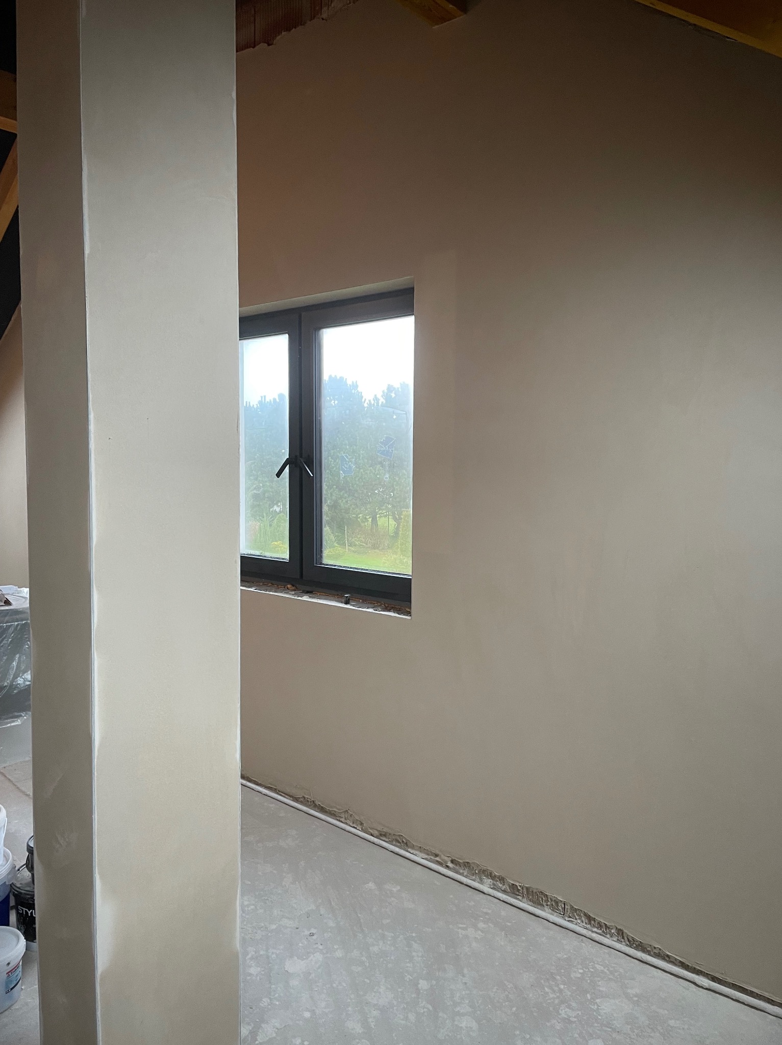 Wnętrze po tynkowaniu maszynowym: gładkie ściany w jasnym odcieniu, okno z widokiem na zieleń, fragment kolumny i podłoga z resztkami zaprawy.