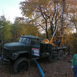 Ciężarówka z wiertnicą na tle jesiennego lasu, z niebieską rurą wychodzącą spod pojazdu i mężczyzną w zielonej bluzie patrzącym w obiektyw.