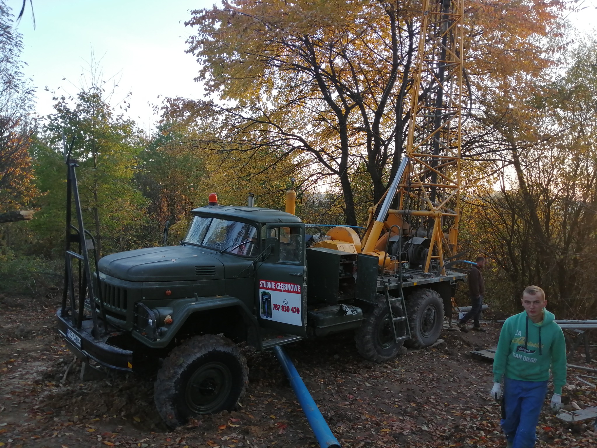 Ciężarówka z wiertnicą na tle jesiennego lasu, z niebieską rurą wychodzącą spod pojazdu i mężczyzną w zielonej bluzie patrzącym w obiektyw.