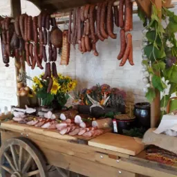 Stoisko w stylu rustykalnym z wędlinami, kiełbasami i dekoracjami roślinnymi, w tle biała ściana z cegły.