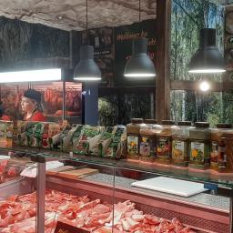 Firma Tołłoczko - Wnętrze sklepu mięsnego z dwoma pracownicami za ladą, ekspozycja różnych rodzajów surowego mięsa w chłodzonych gablotach, przyprawy w słoikach na górnej półce, dekoracja ściany z motywem leśnym...