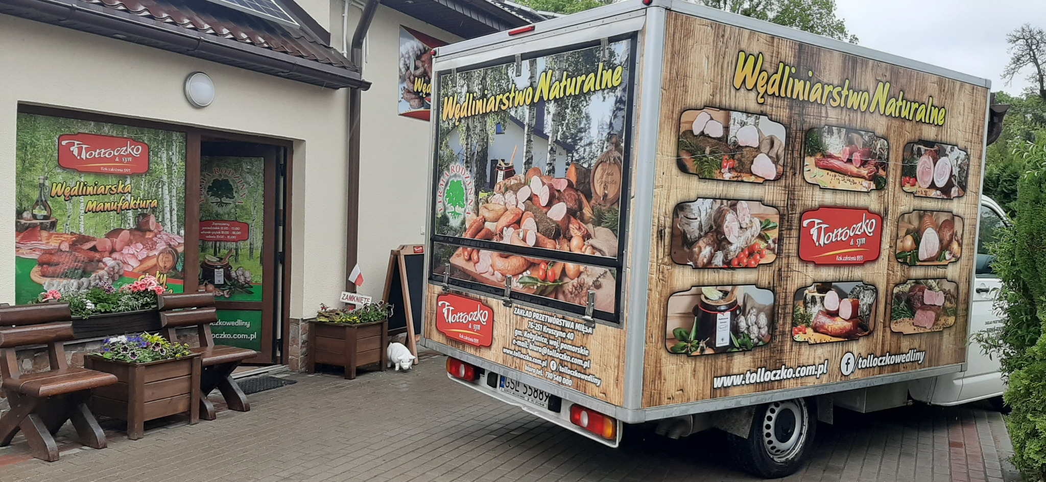 Firma Tottoczko & syn: widok na budynek zakładu z przyczepą reklamową prezentującą wędliniarstwo naturalne, z drewnianymi ławkami i kwiatami na pierwszym planie, oraz białą figurką świnki obok...