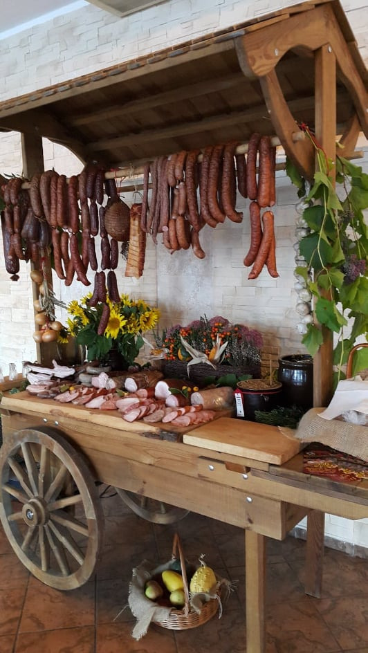 Stoisko w stylu rustykalnym z wędlinami, kiełbasami i dekoracjami roślinnymi, w tle biała ściana z cegły.