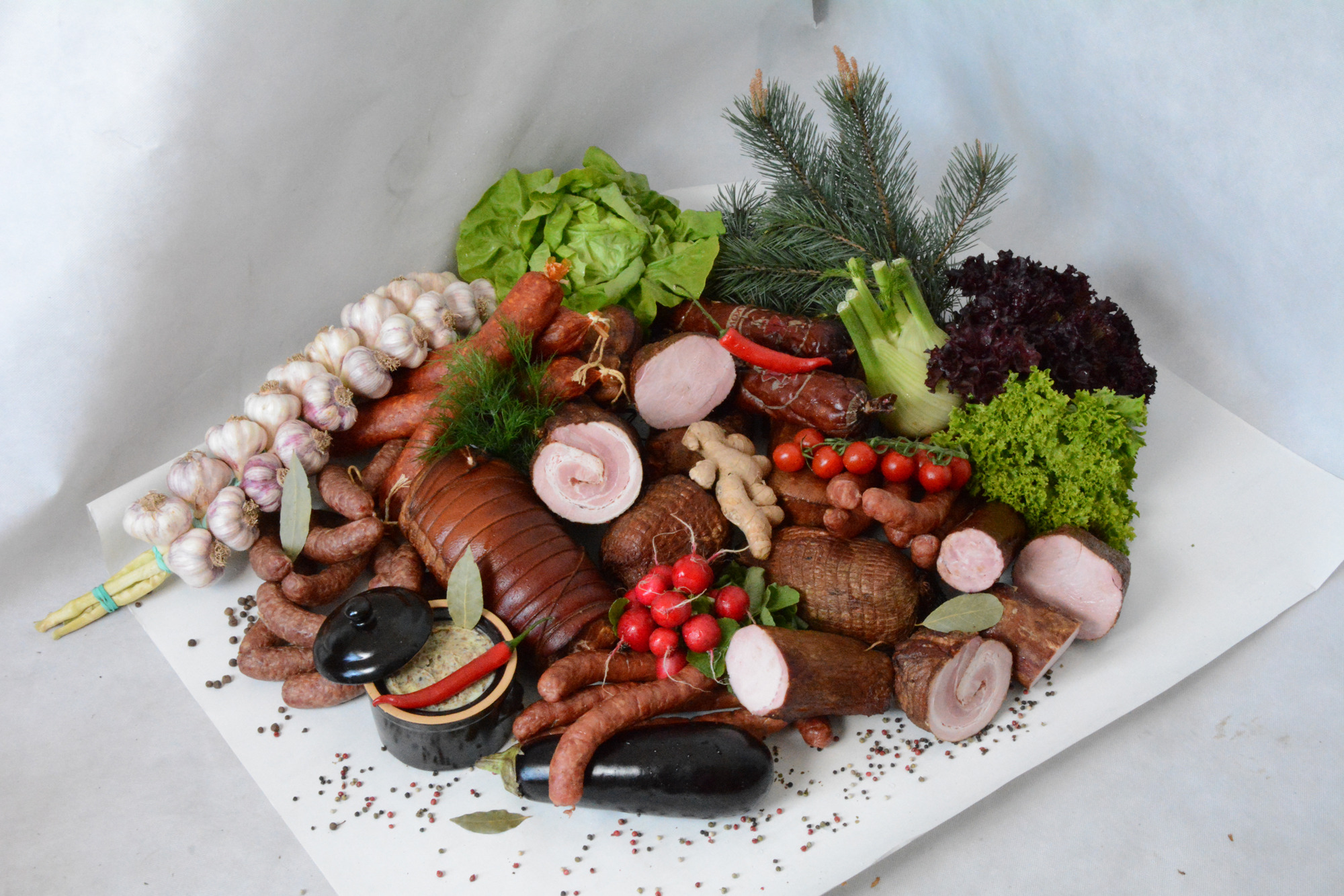 Bogaty asortyment wędlin i kiełbas, z czosnkiem, warzywami i przyprawami, ułożony na białym tle, z gałązką świerku, tworzący kompozycję delikatesową.
