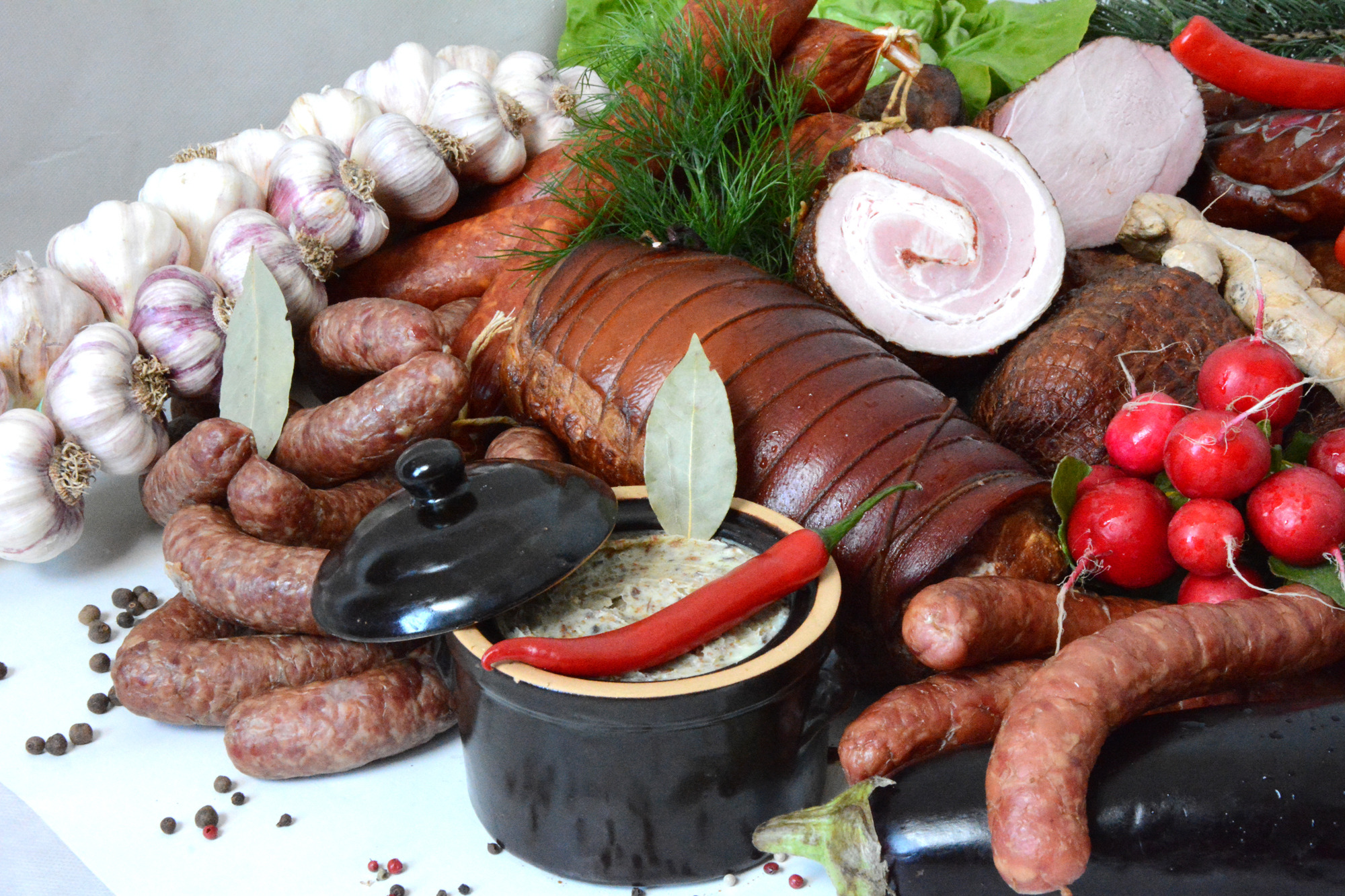 Kompozycja wędlin, kiełbas, boczku rolowanego, czosnku, rzodkiewek i smalcu z papryczką w czarnym garnku, udekorowana liściem laurowym i koperkiem.