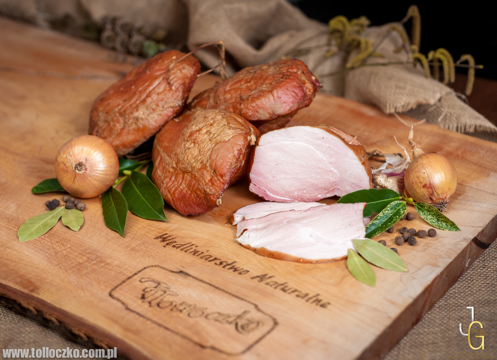 Wędzone wyroby na drewnianej desce z cebulą, liśćmi laurowymi i pieprzem, logo firmy rzeźniczej wygrawerowane na desce.