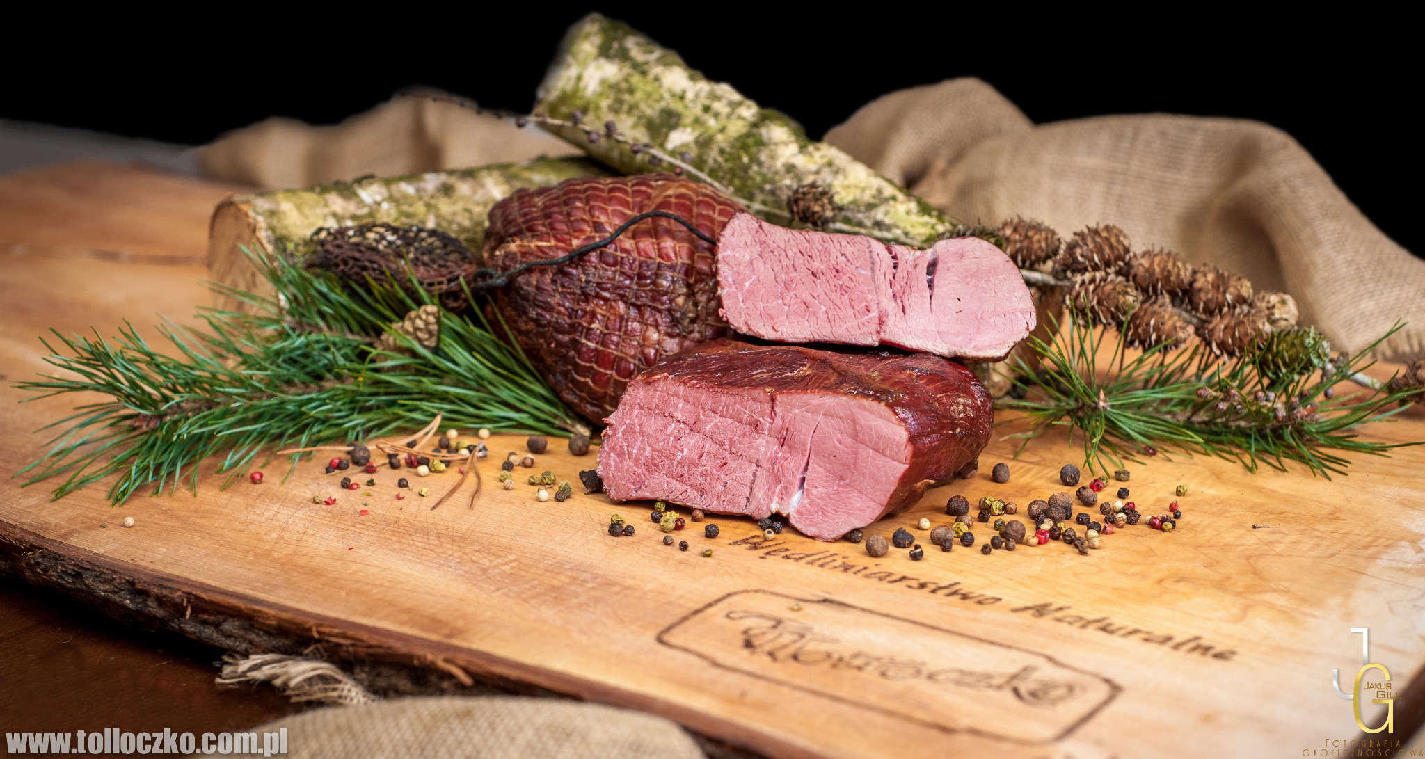 Wędzona polędwica przekrojona na drewnianej desce z napisem 'Wędliniarstwo Naturalne', ozdobiona gałązkami sosny, szyszkami, pieprzem ziarnistym i korą brzozy w tle.