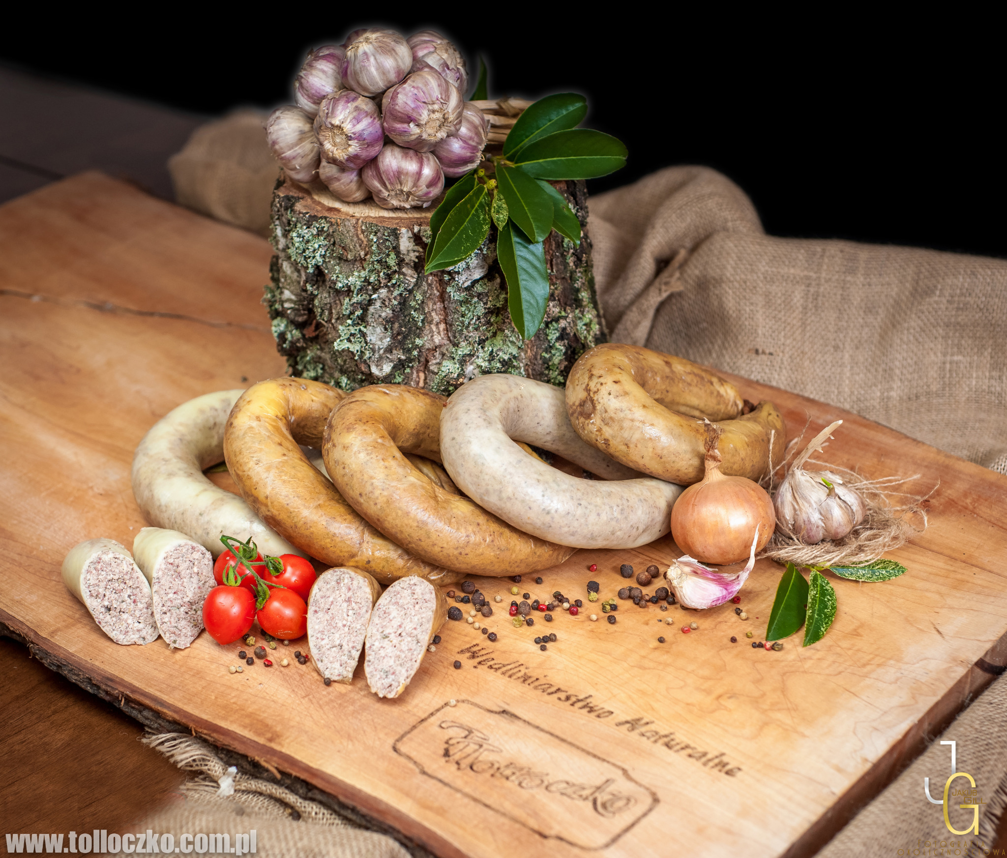 Obróbka wędliniarska: Różne rodzaje kiełbas w kształcie okręgów, w tym biała, ułożone na drewnianej desce z napisem 'Wędliniarstwo Naturalne Tołłoczko', obok pomidory koktajlowe na gałązce, cebula,...