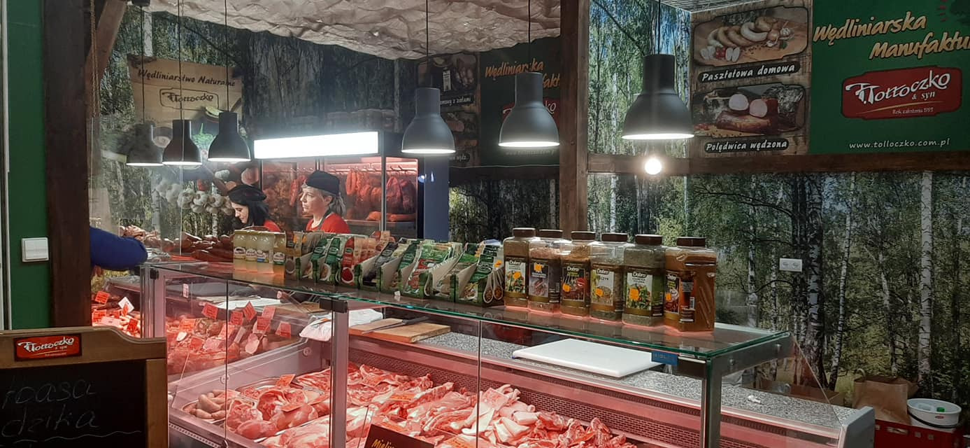 Wnętrze sklepu mięsnego z dwoma pracownicami za ladą, ekspozycja różnych rodzajów surowego mięsa w chłodzonych gablotach, przyprawy w słoikach na górnej półce, dekoracja ściany z motywem leśnym...