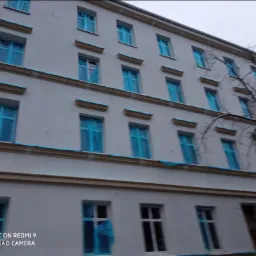 Wielopiętrowy budynek z jasną elewacją, okna zabezpieczone folią ochronną, widoczne elementy dekoracyjne na fasadzie, pochmurne niebo w tle.