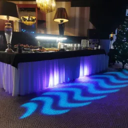 Stół cateringowy z czarnym obrusem i białymi falbanami, oświetlony niebieskim światłem w kształcie fal, w tle choinka i zastawa stołowa, realizacja w Zielonce.