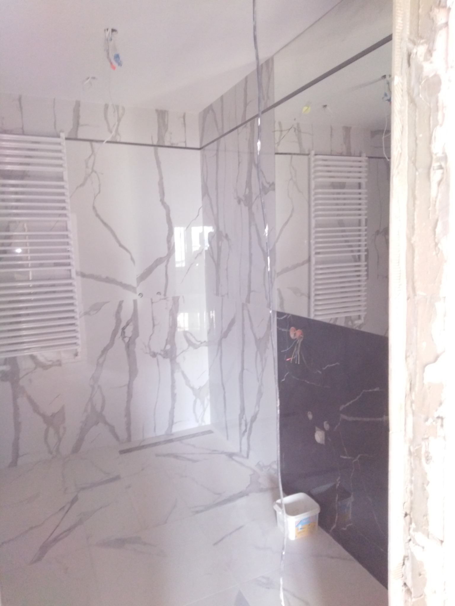 Wnętrze łazienki w trakcie prac wykończeniowych: białe marmurowe płytki na ścianach i podłodze, grzejnik drabinkowy, fragment czarnej ściany z instalacją elektryczną, widoczne przewody i puszka...