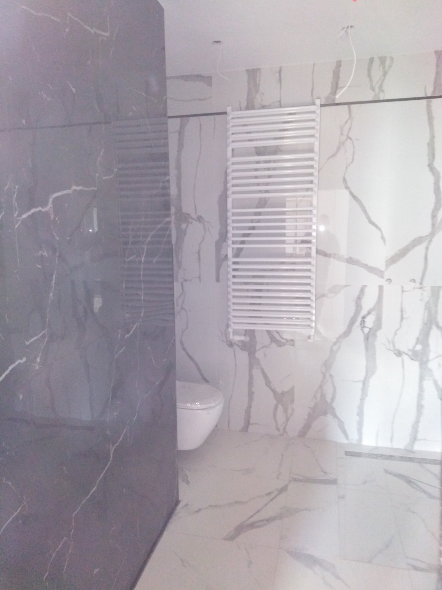 Nowoczesna łazienka z marmurowymi płytkami w odcieniach szarości i bieli, widoczny biały grzejnik drabinkowy i fragment toalety.