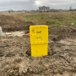 Żółta skrzynka gazowa PSG w ziemi na placu budowy, widoczny fragment rury, w tle dom jednorodzinny i pochmurne niebo.