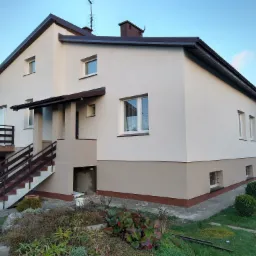 Dwukondygnacyjny dom jednorodzinny z nową, dwukolorową elewacją w odcieniach beżu i brązu, widok z ogrodu na front budynku z drewnianymi schodami i balkonem.