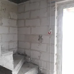 Surowa ściana z betonu komórkowego w trakcie remontu, widoczne białe przewody elektryczne prowadzone po powierzchni, puszka instalacyjna z pomarańczowym wnętrzem oraz fragment betonowych schodów.