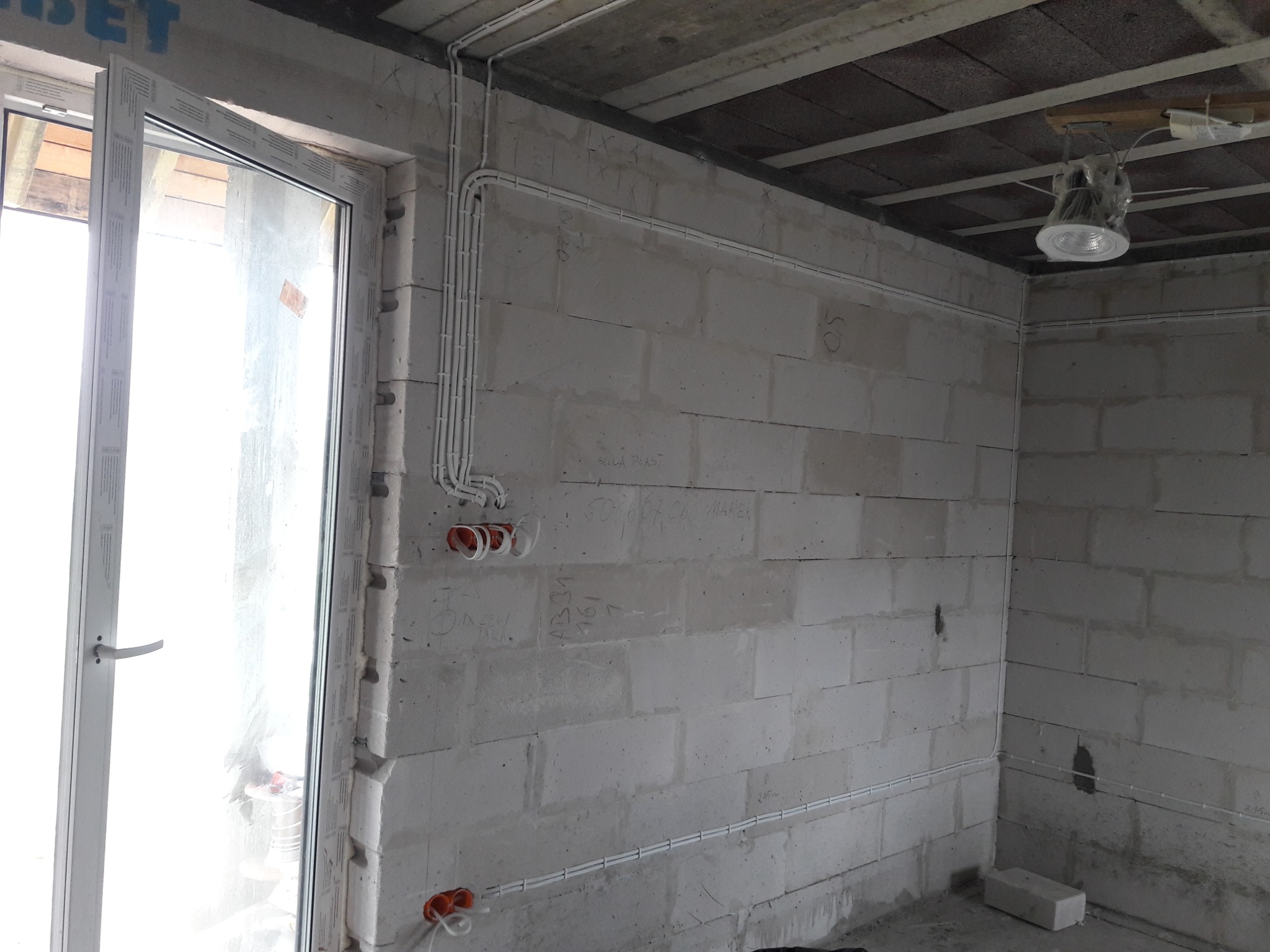 Surowy pokój z białych bloczków, widoczne białe kable elektryczne ułożone w listwach na ścianach, puszki pod gniazdka i lampa sufitowa w folii ochronnej, okno z widokiem na zewnątrz.