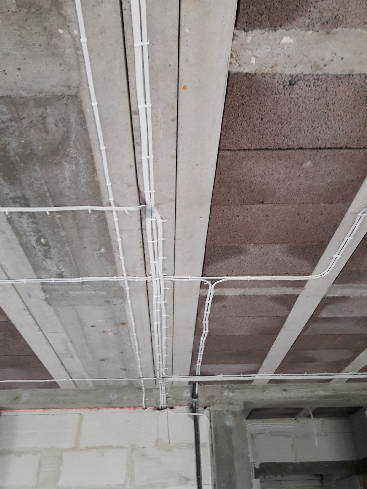 Białe kable elektryczne prowadzone po suficie z betonu komórkowego i pustaków, zabezpieczone uchwytami, widoczne elementy konstrukcyjne budynku w surowym stanie.