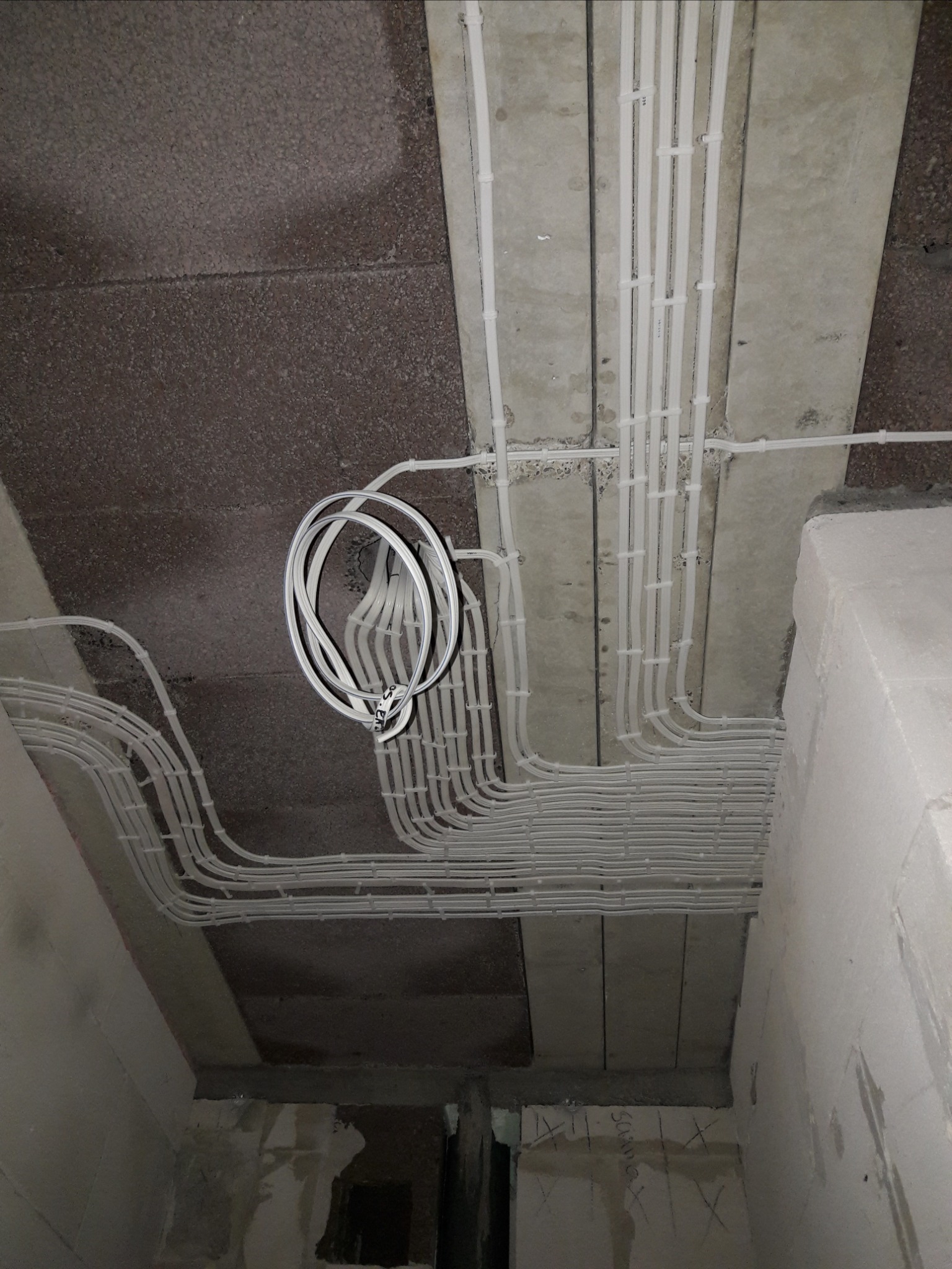 Białe rurki instalacji elektrycznej ułożone równolegle pod betonowym stropem, fragment zwiniętej rury zwisa, widoczne mocowania kabli do sufitu.