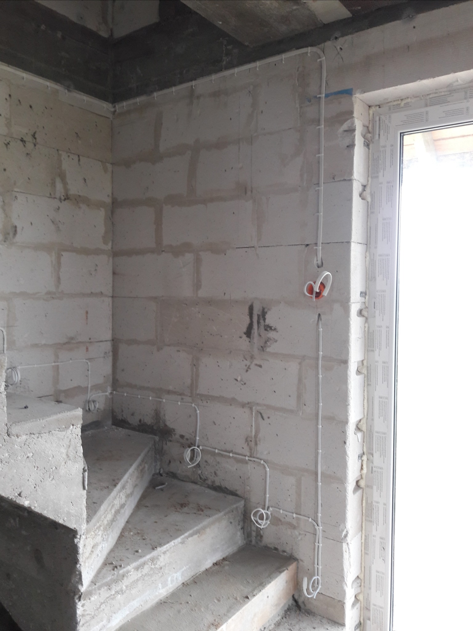 Surowa ściana z betonu komórkowego w trakcie remontu, widoczne białe przewody elektryczne prowadzone po powierzchni, puszka instalacyjna z pomarańczowym wnętrzem oraz fragment betonowych schodów.