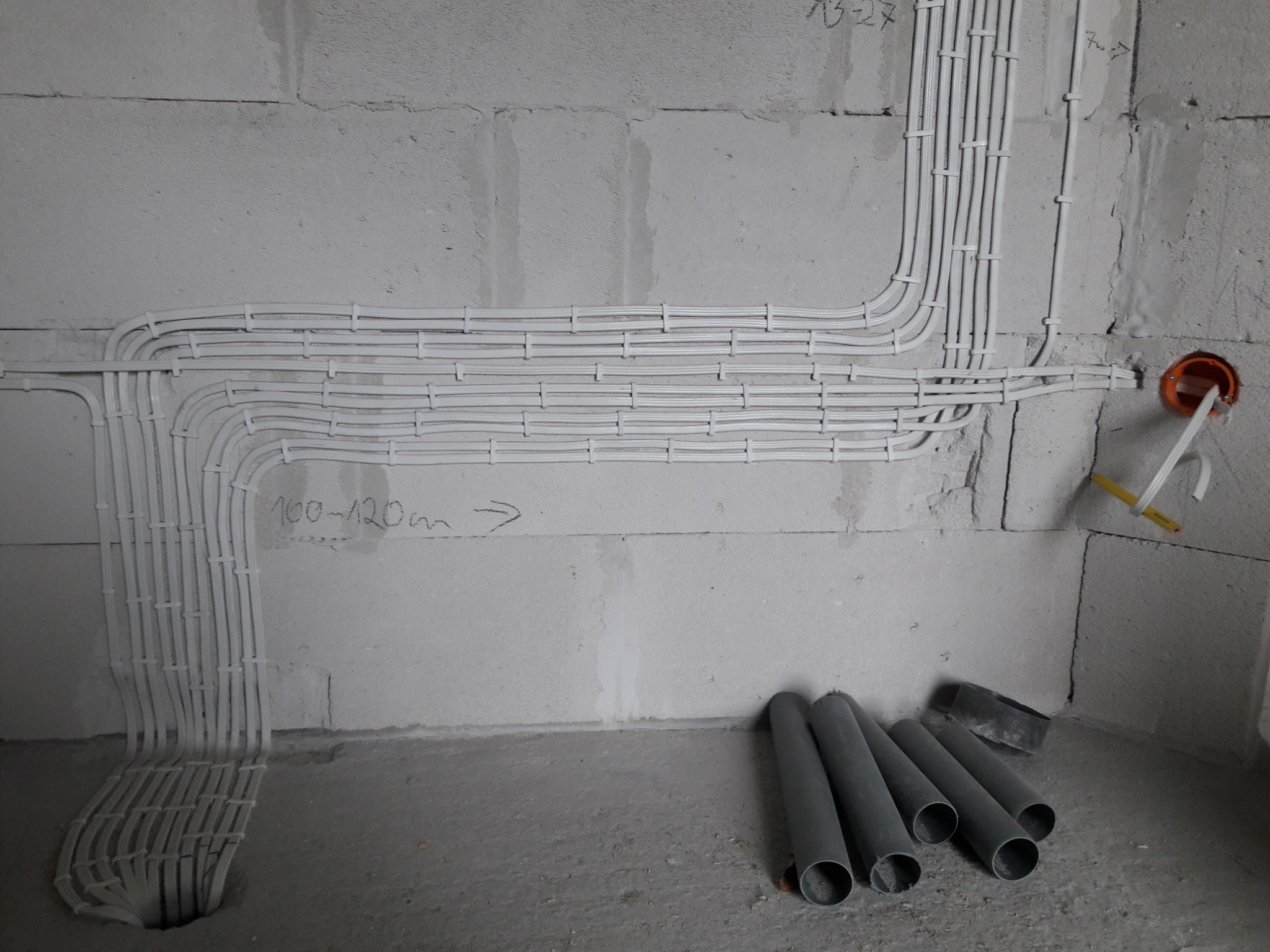 Białe kable elektryczne ułożone w peszlach, przymocowane do surowej ściany z betonu komórkowego, widoczne szare rury PCV na podłodze i pomarańczowa puszka instalacyjna w ścianie.