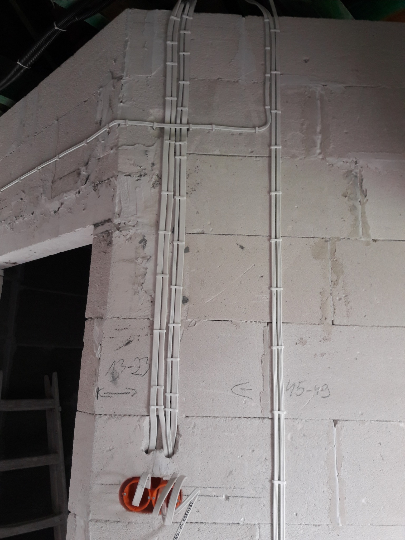 Białe przewody elektryczne prowadzone pionowo po surowej ścianie z betonu komórkowego, zabezpieczone opaskami zaciskowymi, z widoczną pomarańczową puszką instalacyjną i notatkami ołówkiem na ścianie.