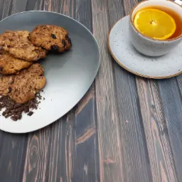 Owsiane ciasteczka z kawałkami czekolady na szarym talerzu obok filiżanki herbaty z plasterkiem pomarańczy na drewnianym tle.