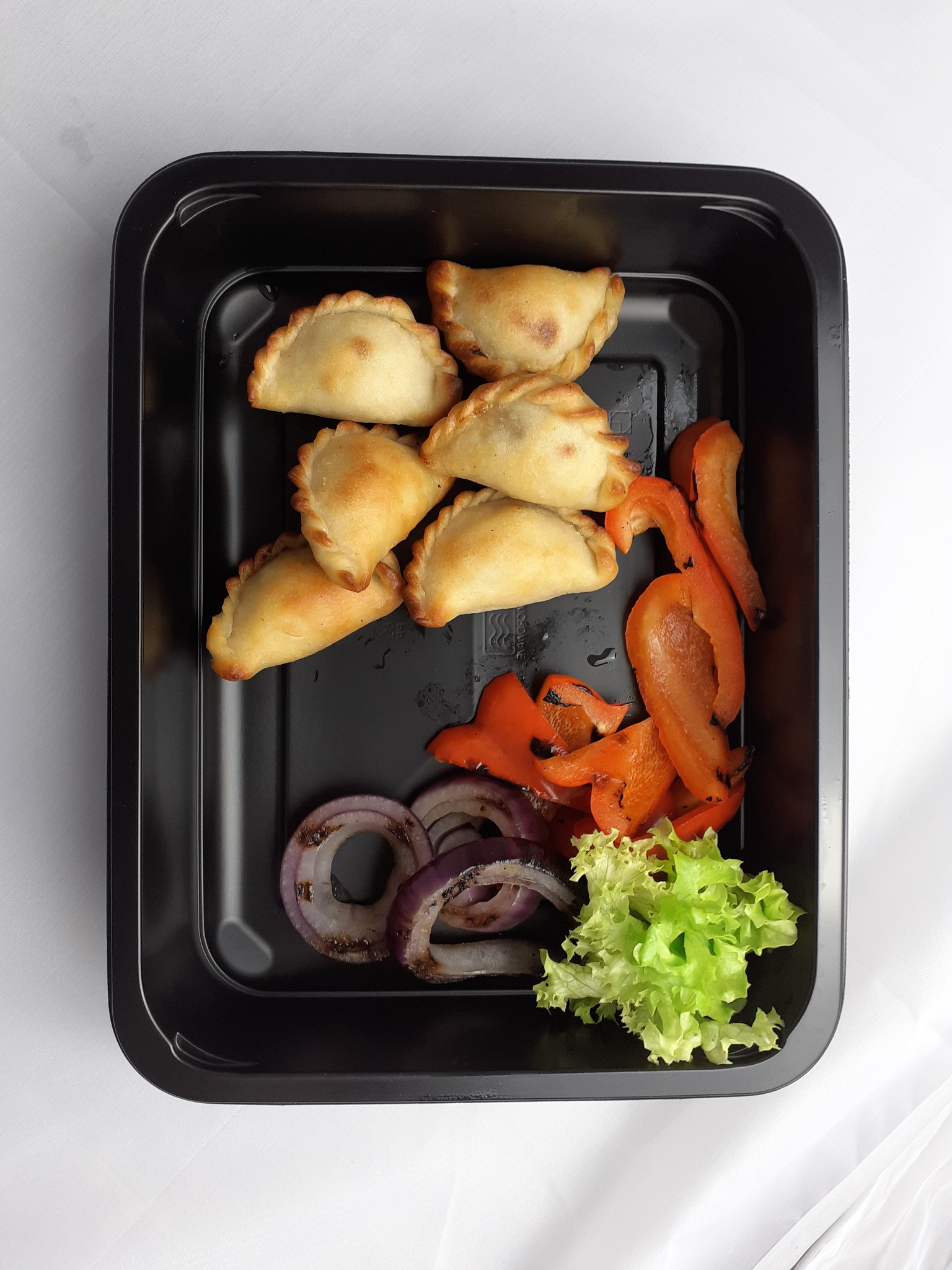 Czarny pojemnik z pieczonymi pierożkami, grillowaną papryką, krążkami czerwonej cebuli i sałatą karbowaną na białym tle.