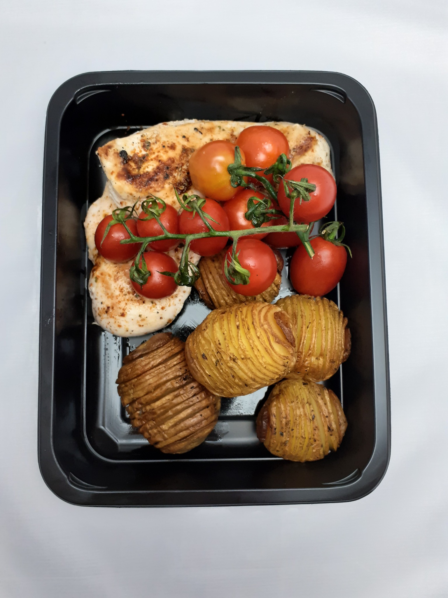 Gotowy posiłek w czarnym pojemniku: grillowana pierś z kurczaka, pieczone ziemniaki hasselback i gałązka pomidorków koktajlowych.