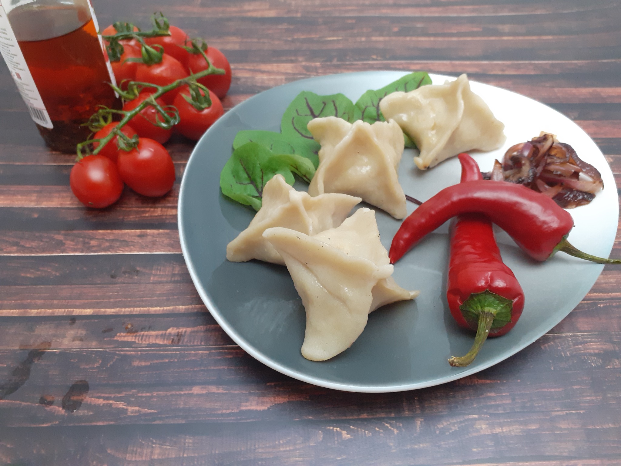 Szare talerz z gotowanymi pierogami w kształcie trójkątów, czerwoną papryką, smażoną cebulą i sałatą, obok butelka oleju i pomidorki koktajlowe na drewnianym stole.
