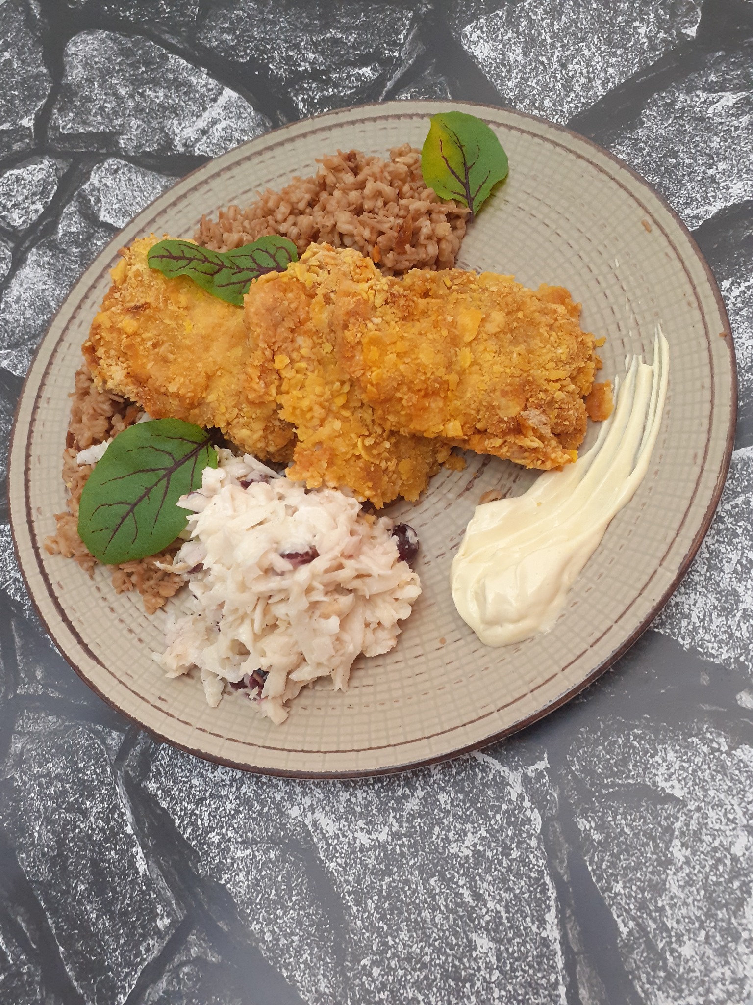 Dwa kawałki panierowanej ryby na talerzu z kaszą gryczaną, surówką colesław i kleksem majonezu, udekorowane listkami.