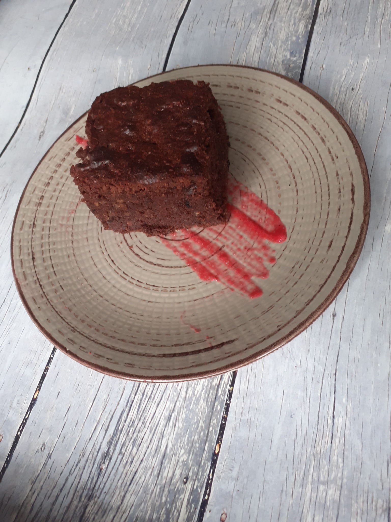 Kwadratowy kawałek ciemnego ciasta czekoladowego na talerzu z beżowym, koncentrycznym wzorem i czerwoną dekoracją z sosu owocowego, ułożony na tle z desek o szarym odcieniu.