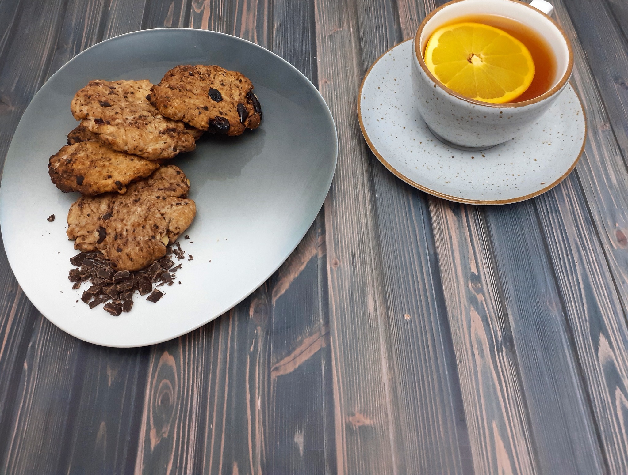 Owsiane ciasteczka z kawałkami czekolady na szarym talerzu obok filiżanki herbaty z plasterkiem pomarańczy na drewnianym tle.